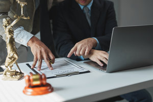 Two lawyers are discussing contract details, pointing at specific clauses on a clipboard, while using a laptop for legal research and documentation in their office
