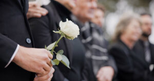 AdobeStock 2030273286 Hands, rose and a person at a funeral in a cemetery in grief while mourning loss at a memorial service. Death, flower and an adult in a suit at a graveyard in a crowd for an outdoor burial closeup